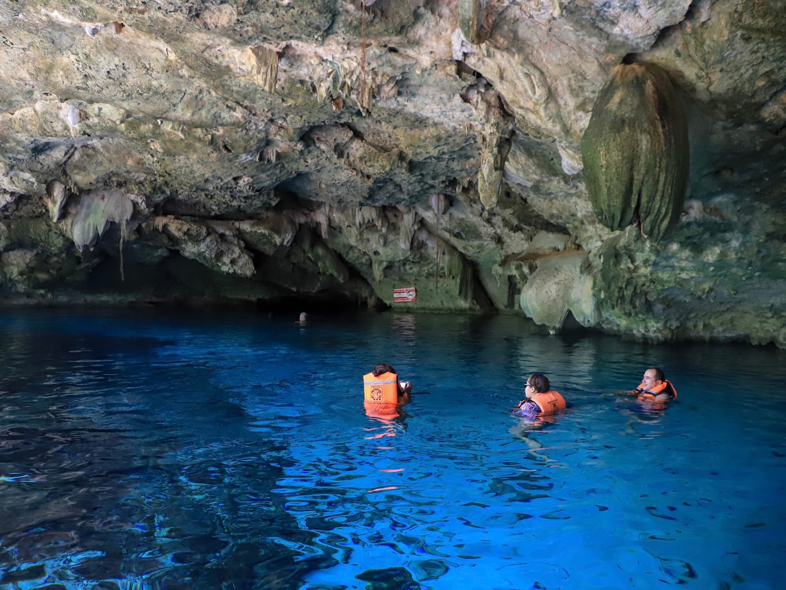 Three guests in life jackets floating beneath a large stalactite in Cenote Dos Ojos