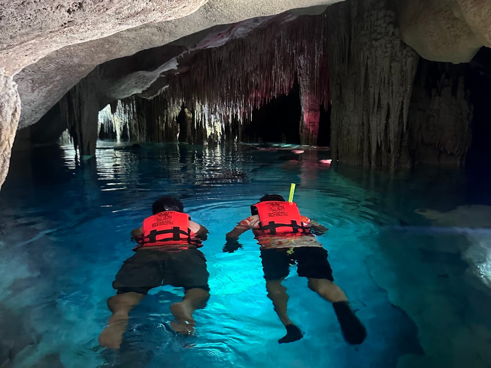 Two snorkelers in life jackets glide along a lit underground river between stalactites, Cenote Los Monos